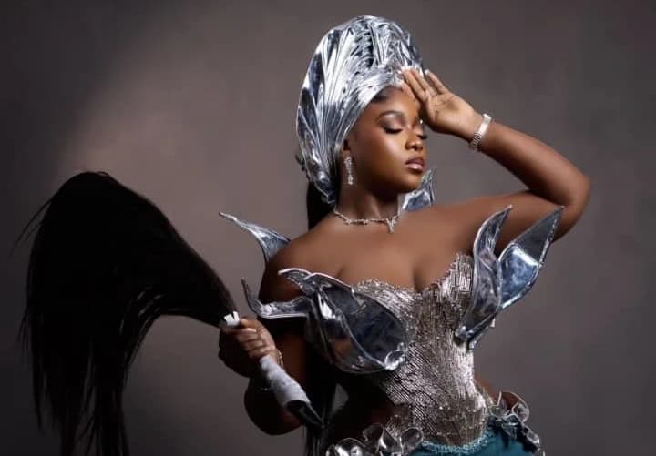 A woman is posing in a striking costume, likely for a fashion show. She is wearing a silver headpiece, a shimmering silver top with decorative accents, and holding a black object resembling a horsetail. The image is well-lit and professionally styled.