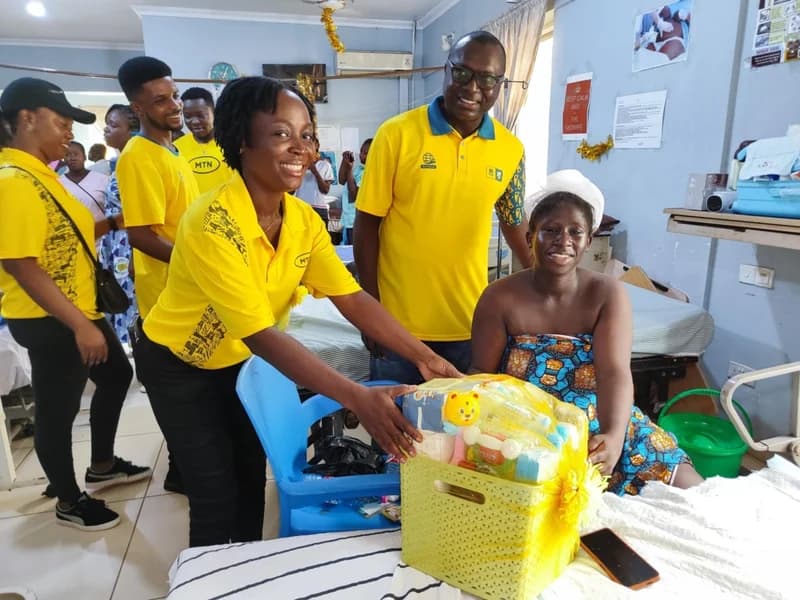 MTN Ghana staff members in their signature yellow shirts are seen presenting a large gift hamper to a new mother in a hospital ward. The woman is seated on her bed and smiling warmly as she receives the donation, which is part of a seasonal charity initiative in Takoradi.