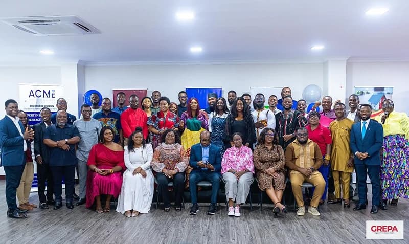 A group photograph of several dozen real estate professionals gathered indoors for a formal event. The attendees, dressed in a variety of business and traditional Ghanaian attire, are posed together in front of corporate banners, including one for the ACME Institute of Real Estate.