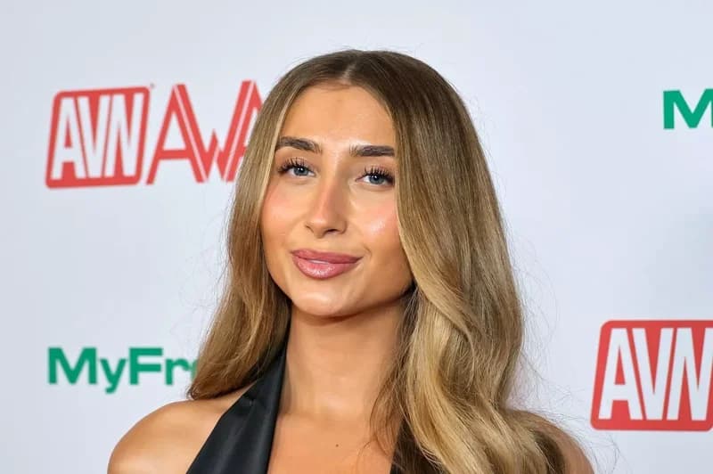 A professional portrait of a woman with long, light-brown hair posing in front of a white media backdrop. She is wearing a black halter-style top and smiling slightly, with the logos for the AVN Awards visible behind her.