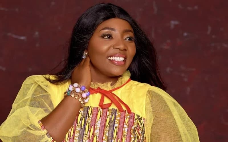 A portrait of Ghanaian singer Afia Ayiwah, who is smiling and looking slightly off-camera. She is wearing a vibrant yellow outfit with sheer sleeves and a patterned bodice, accessorized with layered beaded bracelets.