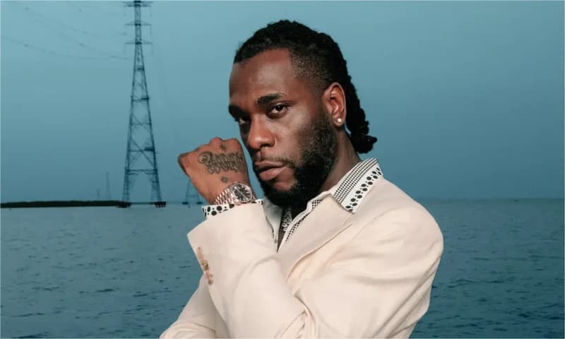 A portrait of Nigerian Afrobeat star Burna Boy wearing a light-colored suit jacket and a luxury watch. He is standing in front of a body of water with a large power transmission tower visible in the background under a dusky blue sky.