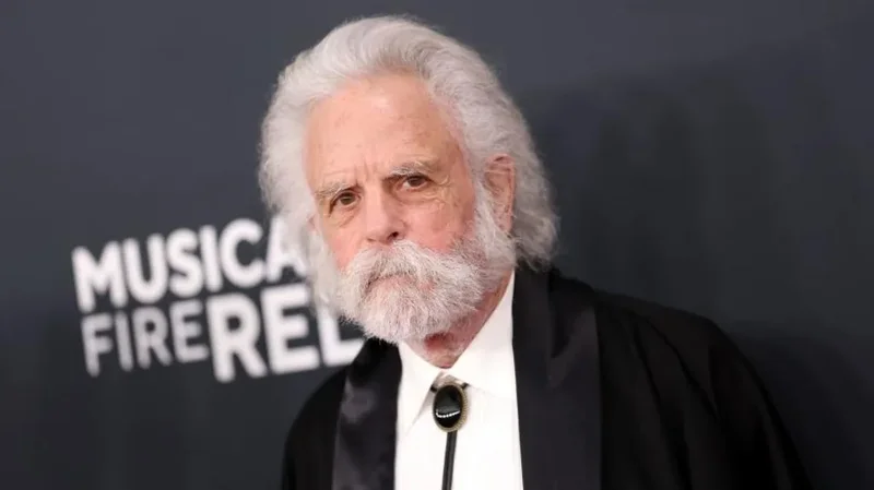 A professional portrait of Bob Weir, the co-founder of the Grateful Dead, featuring his signature long white hair and thick white beard. He is seen wearing a white dress shirt with a black bolo tie under a dark jacket against a promotional backdrop.