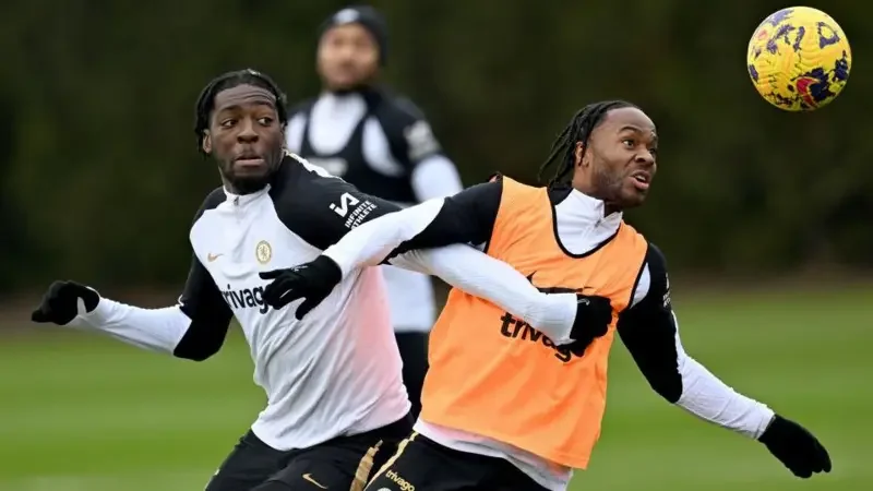Chelsea players Raheem Sterling and Axel Disasi are pictured during a training session on a grass pitch. Sterling, wearing an orange bib, and Disasi, in a white training top, are shown in an active drill with a yellow football suspended in the air. The image captures the intensity of professional training as both players look focused on the ball.