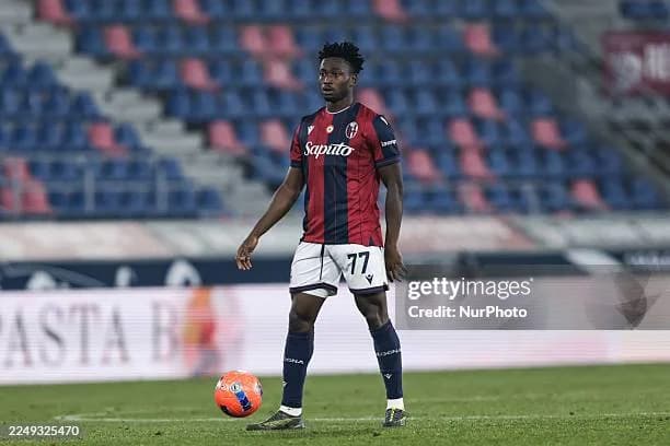 This image shows Ibrahim Sulemana, a Ghanaian footballer, in his Bologna FC jersey during a match. He is on the field, with a soccer ball at his feet, focused on the game. The background features stadium seating.