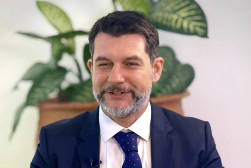 A portrait of Leandro Medina, the IMF Resident Representative to Ghana, appearing to speak during an interview or formal briefing. He is wearing a professional dark suit and a blue tie with red polka dots, with a green potted plant visible in the background.