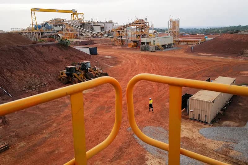 An expansive aerial view of an industrial mining site, featuring large-scale processing infrastructure such as conveyor belts and a yellow gantry crane. Two heavy-duty front loaders are parked on the reddish-brown earth, while a lone worker in high-visibility safety gear walks through the middle ground.
