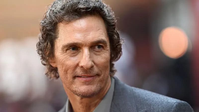 A close-up portrait of actor Matthew McConaughey looking slightly to the side with a gentle expression. He has wavy, salt-and-pepper hair and is wearing a professional grey suit jacket over a light green shirt against a blurred background.