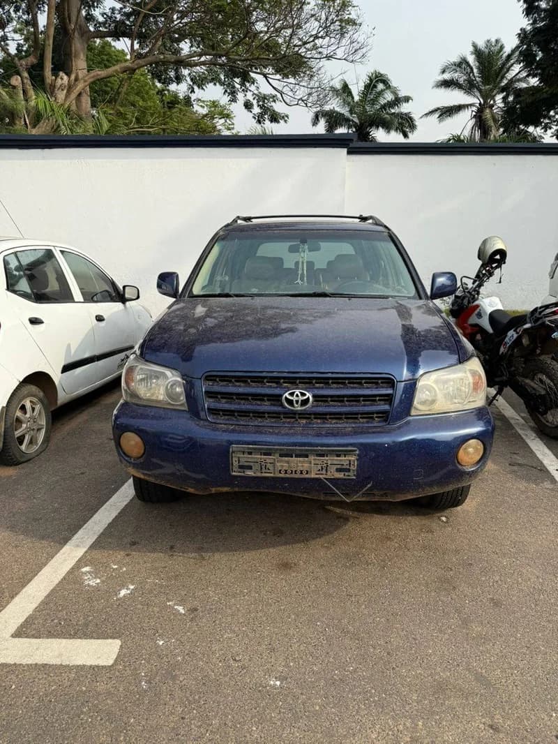 A blue Toyota Highlander is parked in a designated parking space, facing the camera directly. The vehicle appears slightly dusty, and its license plate is obscured or missing. A white car is parked to the left, and a police motorcycle is to the right.