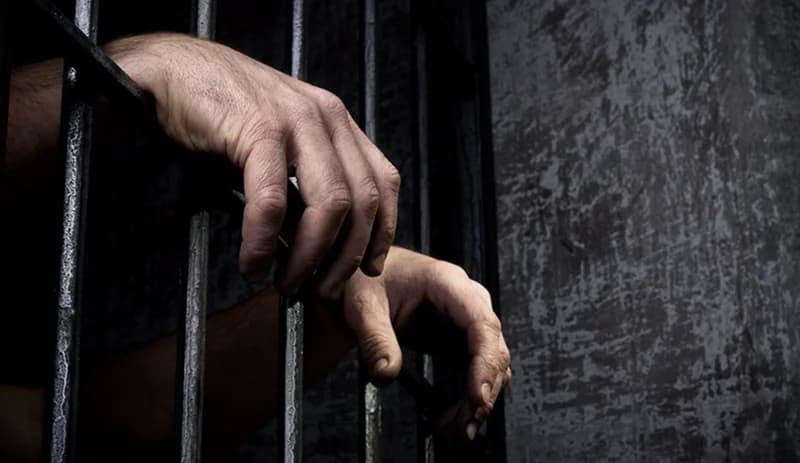 A close-up photograph showing a pair of hands gripping dark metal prison bars. The background consists of a weathered grey wall, creating a somber and dramatic atmosphere representative of incarceration.
