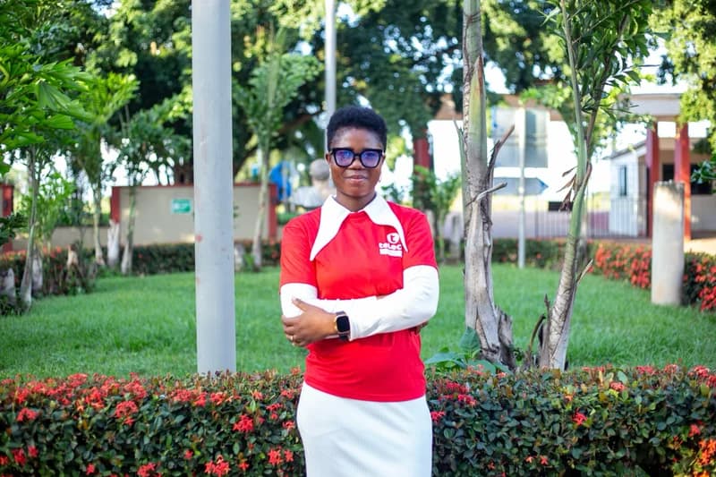 A woman stands confidently with her arms crossed in a lush outdoor garden setting, wearing a red Telecel branded t-shirt and glasses. She is positioned centrally in the frame, surrounded by green grass, trees, and red flowering hedges.