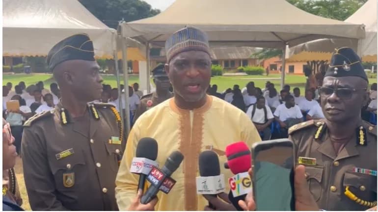 The image shows a man in a yellow traditional smock and matching kufi hat addressing the media during an outdoor event. He is flanked by two high-ranking officials in brown uniforms, likely from the Ghana Prisons Service, while numerous recruits in white shirts are visible in the background under canopies.
