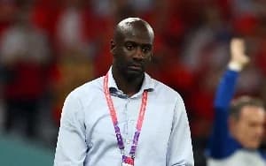 The image shows Otto Addo, the former coach of the Ghana Black Stars, during what appears to be a match. He is wearing a light blue shirt and a lanyard. The background features a blurred crowd in red and white.