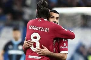 The image shows two Liverpool football players embracing on the field. One player, wearing the number 8 jersey with the name "SZOBOSZLAI" on the back, is hugging Mohamed Salah, who is facing the camera. The background shows a blurry view of the stadium and other players.