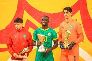 The image shows three soccer players posing with trophies. Sadio Mane is holding a golden trophy, and the other two players are holding smaller awards, one appears to be a golden shoe. They are all wearing athletic wear, standing in front of a yellow and red background.