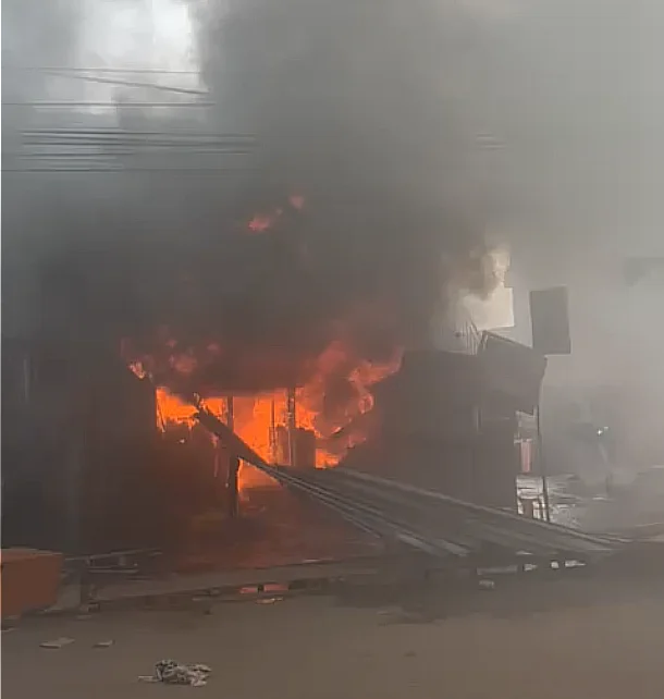 A building identified as shops in Madina is engulfed in intense orange flames with thick, dark grey smoke billowing into the sky. Corrugated metal roofing sheets and other debris are scattered on the ground in front of the burning structure.