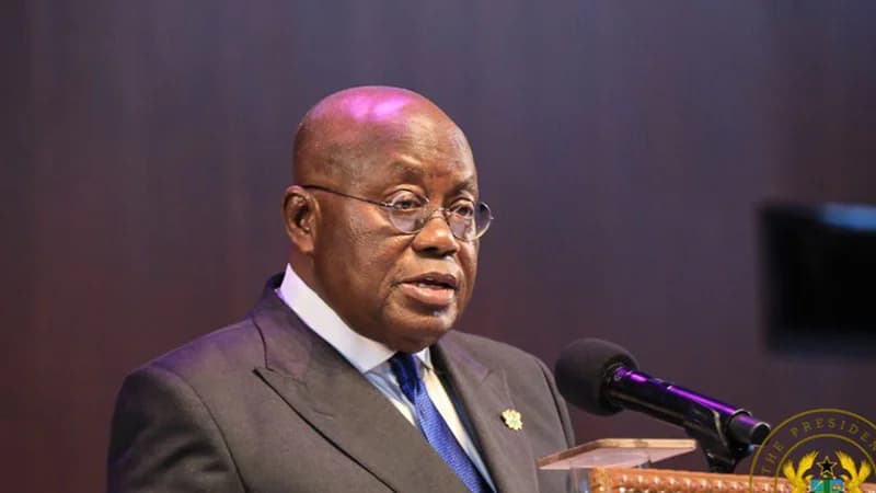 A close-up photograph of Nana Akufo-Addo, the former President of Ghana, speaking at a podium. He is dressed in a formal dark grey suit with a blue tie and his signature round spectacles, positioned in front of a microphone as if delivering a speech.