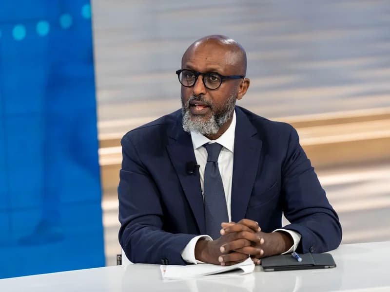 A close-up photograph of Abebe Aemro Selassie, the Director of the African Department at the IMF. He is depicted wearing a dark navy suit and glasses, seated at a desk in what appears to be a media studio environment, speaking while his hands are clasped together.
