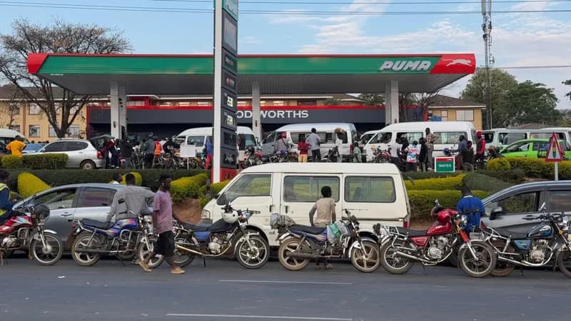 A crowded Puma petrol station is shown with numerous motorcycles and white minibuses queuing for fuel. Motorists and bystanders are seen gathered around the pumps and the forecourt against a backdrop of a 'Foodworths' store and a price display pylon.