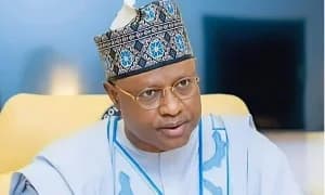 The image shows a portrait of a man wearing glasses and a traditional cap. He is dressed in a light blue shirt and appears to be seated in an office setting, with a blurred background. The lighting is well-balanced, highlighting his features.