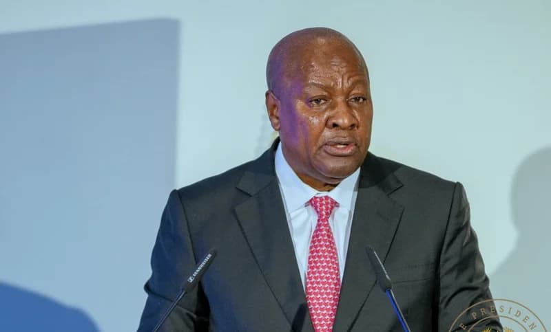 President John Dramani Mahama of Ghana is captured speaking at a formal event, wearing a black suit and a pink patterned tie. He is positioned behind a podium with two Sennheiser microphones visible in the foreground against a neutral background.