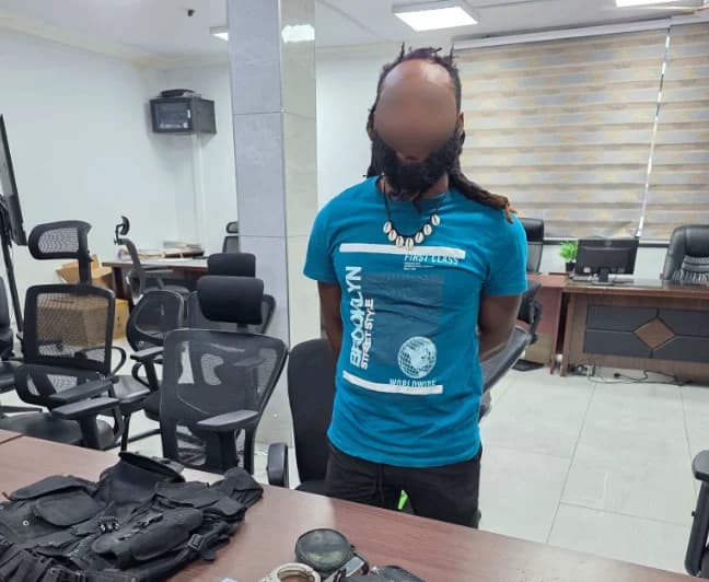 The image shows a man with dreadlocks, whose face has been blurred, standing in what appears to be a police or security office. On a desk in the foreground, law enforcement equipment including a tactical vest, handcuffs, and a firearm are displayed as evidence.