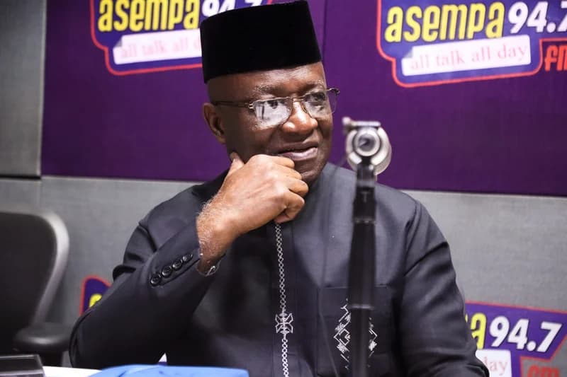 Ghanaian politician Osei Kyei-Mensah-Bonsu is pictured during an interview at the Asempa FM radio studio. He is wearing a black traditional outfit with a matching kufi cap and glasses, appearing thoughtful as he speaks into a studio microphone.