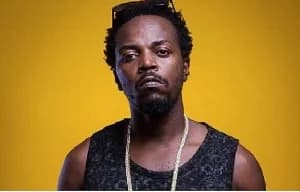 A portrait of Ghanaian musician Kwaw Kese posing against a solid yellow background. He is wearing a black patterned sleeveless shirt, a silver chain, and has sunglasses resting on his head.