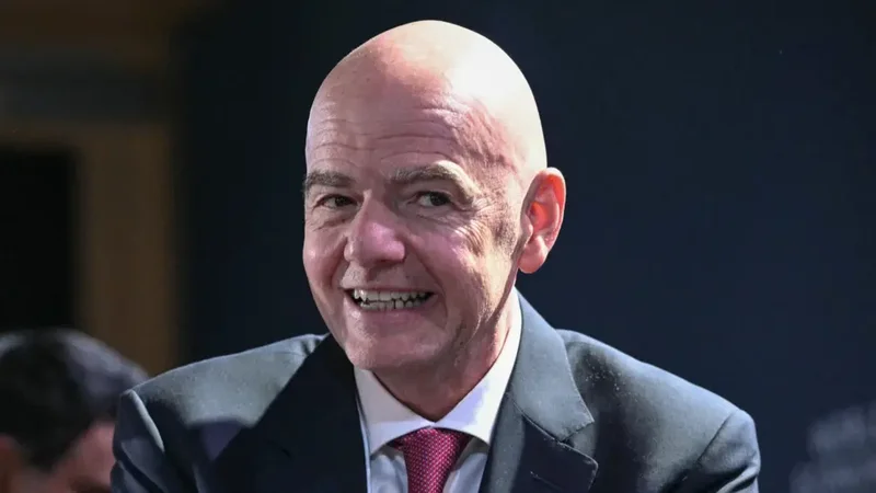 A close-up photograph of FIFA President Gianni Infantino smiling during a public event. He is dressed in a professional dark suit, white shirt, and a burgundy tie against a blurred dark background.