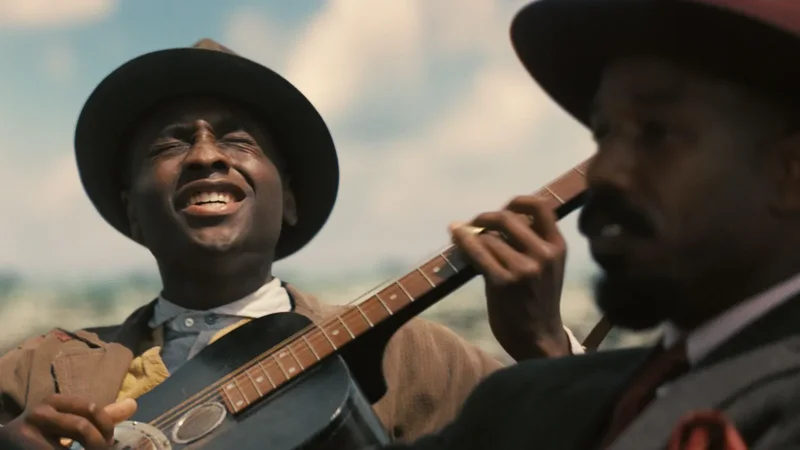 A cinematic still from the movie 'Sinners' featuring two men in vintage attire. One man is shown in sharp focus wearing a fedora while playing an acoustic guitar and singing, while another man in a suit and hat appears blurred in the foreground.