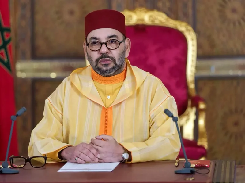 King Mohammed VI of Morocco is seated at a desk wearing traditional attire, including a yellow jellaba and a red fez. He appears to be delivering a formal address or statement, with two microphones positioned in front of him and an ornate background featuring the Moroccan flag.