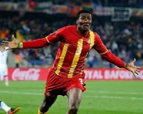 Ghanaian football legend Asamoah Gyan is captured in a triumphant celebration during a match for the Black Stars. He is seen running across the pitch with his arms wide open, wearing the national team's iconic red and yellow striped jersey featuring his signature number three.