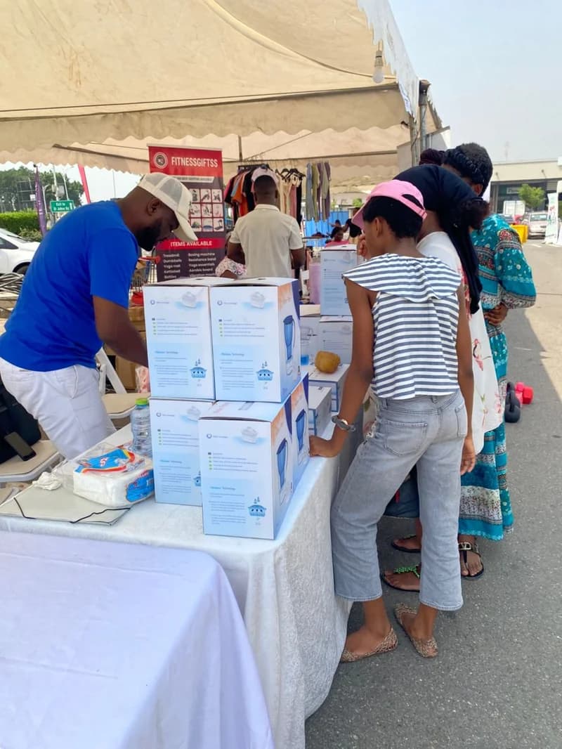 Shoppers browse a variety of household items at an outdoor fair located at the Achimota Retail Centre. A vendor in a blue shirt manages a display of boxed appliances under a white canopy while customers wait to inquire about deals.
