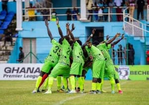 A group of football players in lime green jerseys with black patterns are huddled together with their arms raised in celebration on a grass pitch. The background features stadium seating with spectators and advertising boards prominently displaying 'GHANA Premier League' and the logo for Bechem United FC.