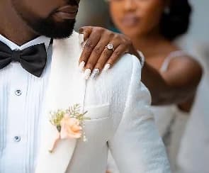 A close-up photograph of a groom wearing a white tuxedo and black bow tie, with a bride's hand resting on his shoulder. The bride is visible but out of focus in the background, and her hand features a prominent diamond engagement ring.
