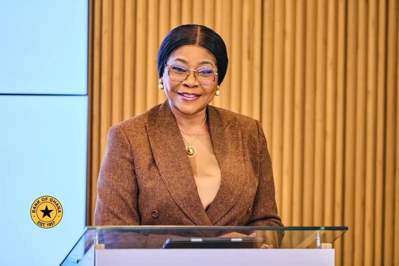 A portrait of Mrs. Elsie Addo Awadzi, the Second Deputy Governor of the Bank of Ghana, smiling while standing behind a glass podium. She is wearing a brown textured blazer and glasses, positioned against a background of light blue panels and vertical wooden slats.