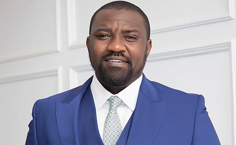 A professional portrait of Ghanaian politician and actor John Dumelo. He is seen wearing a sharp blue suit, a white shirt, and a patterned light-colored tie against a white paneled wall background.