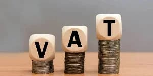 A photograph showing three stacks of coins in increasing height, each topped with a wooden block featuring the letters 'V', 'A', and 'T' respectively. The image serves as a conceptual representation of Value Added Tax and financial thresholds in a business context.