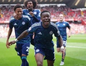 Ghanaian winger Fatawu Issahaku celebrates a goal with his Leicester City teammates during a match. The players are captured in a moment of joy on the field with a blurred crowd in the stadium background.