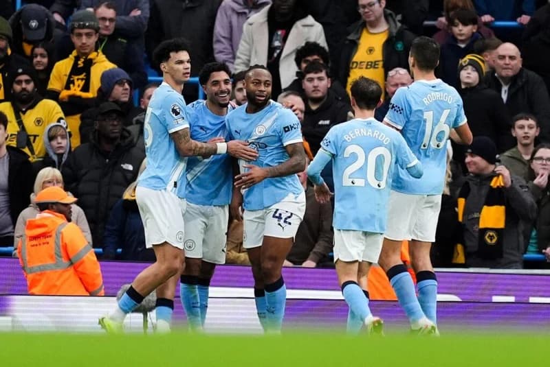 Manchester City players celebrate a goal during an English Premier League match. Bernardo Silva (20) and Rodri (16) are visible among the celebrating group in their iconic light blue home kit, with Wolves supporters in yellow visible in the background stands.