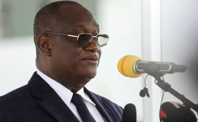 A close-up photograph of a man in a dark formal suit and white shirt, wearing sunglasses and speaking behind a microphone. The image appears to show a high-ranking official during a press briefing or public address, relevant to West African political developments.