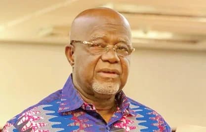 A close-up portrait of Hackman Owusu-Agyeman, a prominent Ghanaian politician and elder of the New Patriotic Party. He is depicted wearing a vibrant blue and pink patterned African print shirt and rectangular glasses.