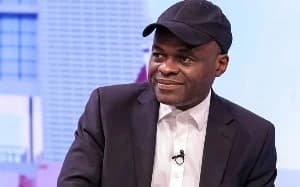 A medium-shot photograph of Ghanaian lawyer Martin Kpebu during a studio interview. He is wearing a dark suit jacket over a white shirt and is notably wearing a black baseball cap with a lapel microphone attached to his clothing.