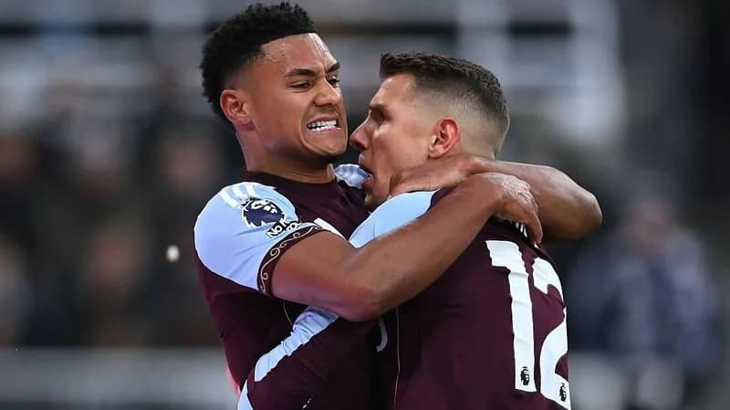 Aston Villa's Ollie Watkins celebrates intensely with a teammate following a goal during a Premier League match. The players are wearing their claret and blue kits, showing a moment of triumph on the pitch against a blurred stadium background.