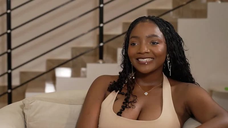 A medium shot of Nigerian singer Simi smiling during what appears to be a sit-down interview. She is wearing a beige halter-neck top and has her hair styled in long braids, seated against a backdrop of a modern staircase.