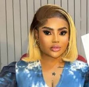 A close-up portrait of Ghanaian media personality Aba Dope, featuring her with a blonde bob hairstyle and full makeup. She is wearing a blue patterned top and gold hoop earrings, posed against a grey slatted background.