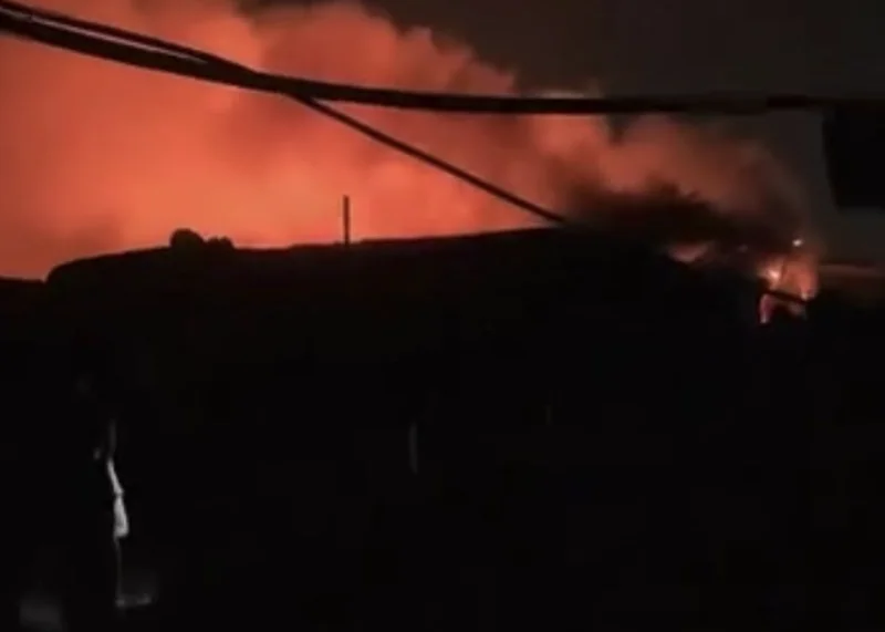 A night scene depicting a massive fire with billowing orange smoke and visible flames rising above the dark silhouettes of market structures. Power lines are visible in the foreground against the glowing sky caused by the blaze.