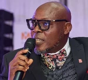 A close-up photograph of Patrice Amegashie, the newly appointed Commercial Manager for GhanaWeb. He is depicted speaking into a handheld microphone while wearing a professional suit and a distinctive patterned bowtie.