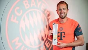 England striker Harry Kane is shown smiling while holding a Bundesliga trophy. He is wearing a red FC Bayern Munich training top in front of a wall featuring an illuminated club logo.