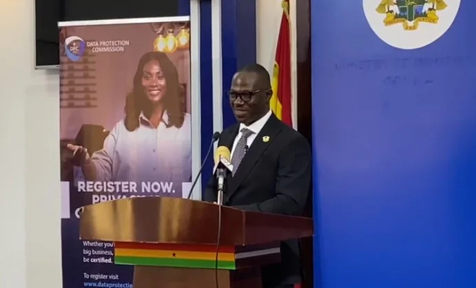 A man in a black suit and glasses delivers a speech at a podium decorated with the colors of the Ghana flag. Behind him, a banner for the Data Protection Commission and a blue backdrop featuring the Ghana Coat of Arms for the Ministry of Information are visible.